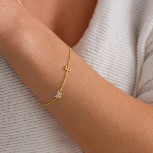 May include: A delicate gold bracelet featuring a butterfly charm and the letter "E". The bracelet is adorned with small, sparkling embellishments. The jewelry is displayed on a person's arm, against a soft, white background.