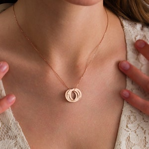 May include: Rose gold necklace featuring three interlocking circles. Each circle has engraved text. The necklace hangs on a delicate chain. The jewelry is displayed on a person wearing a white lace top.