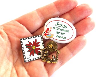May include: A collection of Christmas-themed pins. Includes a square pin with a poinsettia and the year 1997, an oval pin with the text "Jesus is the reason for the Season", and a gold-colored nativity scene pin.