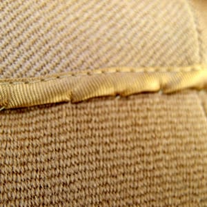 May include: Close-up of a beige fabric ribbon is sewn onto the fabric, creating a clean, finished edge. The image highlights the detail and craftsmanship of the textile.
