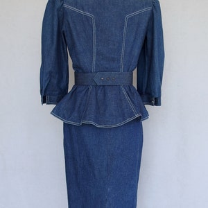 May include: This denim peplum suit is vintage 1980's.  
The jacket has a wide V neckline, silver buttons up the front, puffy 3/4 length sleeves with a single button cuff, and a super flared hemline to create a peplum.  Accented with white topstitching. 

The rigid, obi style belt has faux turquoise  and silver studs.

The unlined  pencil skirt has a slit in the back for wearing ease and a back zipper.