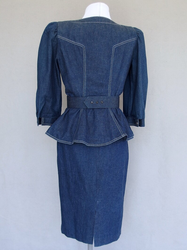 May include: This denim peplum suit is vintage 1980's.  
The jacket has a wide V neckline, silver buttons up the front, puffy 3/4 length sleeves with a single button cuff, and a super flared hemline to create a peplum.  Accented with white topstitching. 

The rigid, obi style belt has faux turquoise  and silver studs.

The unlined  pencil skirt has a slit in the back for wearing ease and a back zipper.
