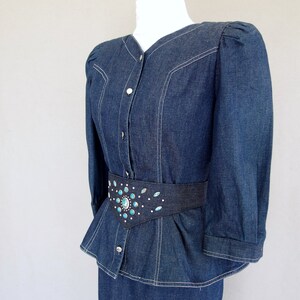 May include: This denim peplum suit is vintage 1980's.  
The jacket has a wide V neckline, silver buttons up the front, puffy 3/4 length sleeves with a single button cuff, and a super flared hemline to create a peplum.  Accented with white topstitching. 

The rigid, obi style belt has faux turquoise  and silver studs.

The unlined  pencil skirt has a slit in the back for wearing ease and a back zipper.