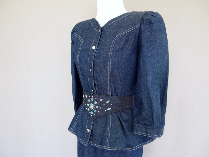 May include: This denim peplum suit is vintage 1980's.  
The jacket has a wide V neckline, silver buttons up the front, puffy 3/4 length sleeves with a single button cuff, and a super flared hemline to create a peplum.  Accented with white topstitching. 

The rigid, obi style belt has faux turquoise  and silver studs.

The unlined  pencil skirt has a slit in the back for wearing ease and a back zipper.