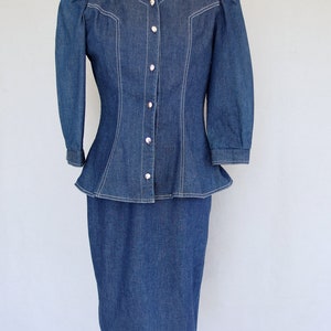May include: This denim peplum suit is vintage 1980's.  
The jacket has a wide V neckline, silver buttons up the front, puffy 3/4 length sleeves with a single button cuff, and a super flared hemline to create a peplum.  Accented with white topstitching. 

The rigid, obi style belt has faux turquoise  and silver studs.

The unlined  pencil skirt has a slit in the back for wearing ease and a back zipper.