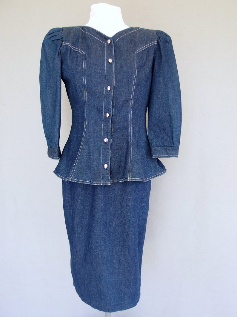 May include: This denim peplum suit is vintage 1980's.  
The jacket has a wide V neckline, silver buttons up the front, puffy 3/4 length sleeves with a single button cuff, and a super flared hemline to create a peplum.  Accented with white topstitching. 

The rigid, obi style belt has faux turquoise  and silver studs.

The unlined  pencil skirt has a slit in the back for wearing ease and a back zipper.