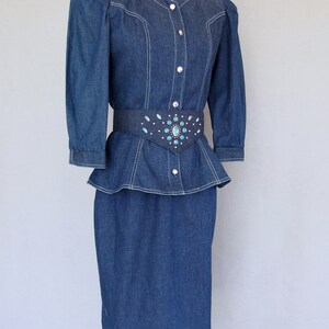 May include: This denim peplum suit is vintage 1980's.  
The jacket has a wide V neckline, silver buttons up the front, puffy 3/4 length sleeves with a single button cuff, and a super flared hemline to create a peplum.  Accented with white topstitching. 

The rigid, obi style belt has faux turquoise  and silver studs.

The unlined  pencil skirt has a slit in the back for wearing ease and a back zipper.
