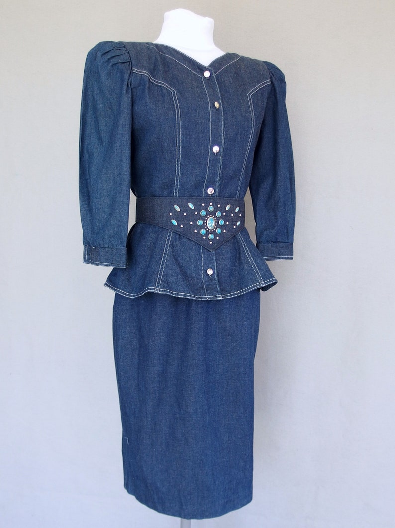May include: This denim peplum suit is vintage 1980's.  
The jacket has a wide V neckline, silver buttons up the front, puffy 3/4 length sleeves with a single button cuff, and a super flared hemline to create a peplum.  Accented with white topstitching. 

The rigid, obi style belt has faux turquoise  and silver studs.

The unlined  pencil skirt has a slit in the back for wearing ease and a back zipper.