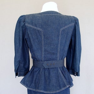 May include: This denim peplum suit is vintage 1980's.  
The jacket has a wide V neckline, silver buttons up the front, puffy 3/4 length sleeves with a single button cuff, and a super flared hemline to create a peplum.  Accented with white topstitching. 

The rigid, obi style belt has faux turquoise  and silver studs.

The unlined  pencil skirt has a slit in the back for wearing ease and a back zipper.
