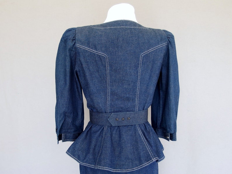May include: This denim peplum suit is vintage 1980's.  
The jacket has a wide V neckline, silver buttons up the front, puffy 3/4 length sleeves with a single button cuff, and a super flared hemline to create a peplum.  Accented with white topstitching. 

The rigid, obi style belt has faux turquoise  and silver studs.

The unlined  pencil skirt has a slit in the back for wearing ease and a back zipper.