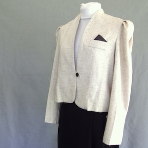 May include: A light beige blazer with a black button closure and a small pocket detail. The blazer has a tailored fit and is paired with a black skirt. The jacket is displayed on a mannequin.