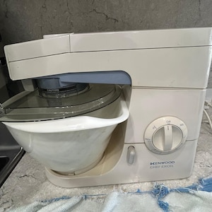 Könnte beinhalten: Eine weiße Kenwood Chef Excel Küchenmaschine mit einer großen weißen Rührschüssel, einem transparenten Spritzschutz und einem runden Drehregler mit den Einstellungen Off, Min und Max. Das Gerät hat ein blaues Akzentteil am Kopf.
