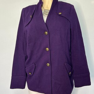 May include: A deep purple jacket featuring a textured herringbone pattern, a stand-up collar, and long sleeves. The front closure includes three gold-toned buttons, with two additional decorative flap pockets at the waist and gold button accents.