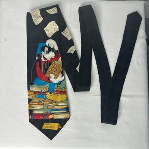 May include: A black necktie featuring a graphic of the cartoon character Goofy wearing a blue graduation cap, reading a book while sitting on a stack of colorful books. The tie includes scattered paper illustrations and a yellow sign reading PROF GOOFY.