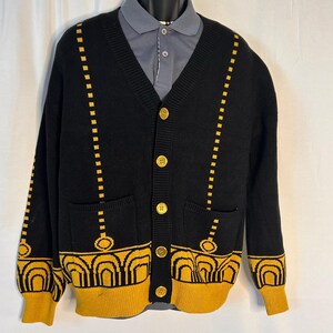 May include: Black cardigan sweater with gold accents. The sweater features a V-neck, button closure, and two front pockets. Gold geometric patterns adorn the cuffs, hem, and vertical lines. The buttons are gold, and the sweater is displayed on a mannequin.