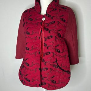 May include: A quilted red jacket featuring a black floral pattern with subtle glitter accents. The jacket has a high collar, three-quarter length sleeves, a single white button closure at the neck, and two thin red ribbons hanging from the button.