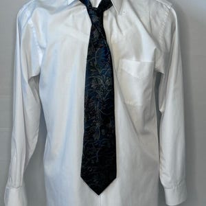 May include: A crisp white long-sleeved button-down shirt paired with a dark navy blue necktie. The tie features an intricate, repeating floral and botanical pattern in shades of light blue, grey, and silver, creating a detailed visual texture.