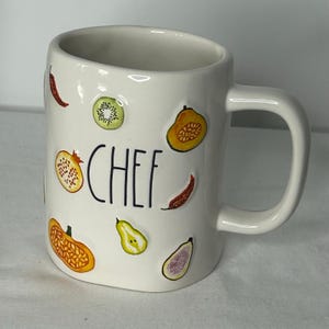 May include: White ceramic mug with the word "CHEF" in black lettering. The mug is decorated with colorful illustrations of fruits and vegetables, including kiwi, pomegranate, and chili peppers. The mug has a curved handle.