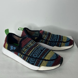 May include: Pair of slip-on sneakers with a colorful woven pattern in blue, red, yellow, and black. The shoes have white soles and a green accent on the heel.