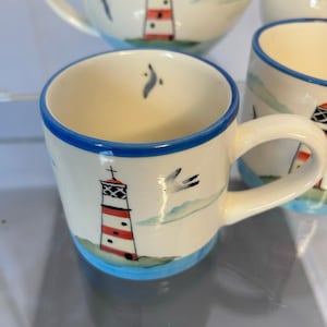 May include: Cream-colored ceramic mug with a blue rim and base, featuring a hand-painted lighthouse design in red and white stripes. The mug also has a seagull and cloud design. Part of a matching set.