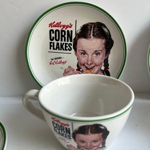 Op de afbeelding: Een witte keramische kom- en bordenset met het Kellogg's Corn Flakes-logo en een vintage illustratie van een meisje dat ontbijtgranen eet. Het bord heeft een groene rand en de kom heeft een handvat.