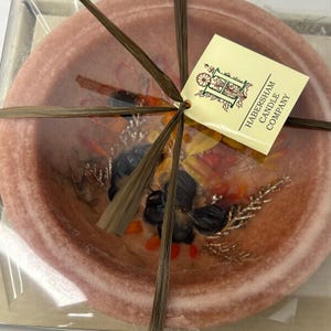 May include: A round, pink, bowl-shaped wax candle embedded with dried dark flowers, orange berries, and gold-toned foliage. The candle is wrapped in thin brown raffia ribbons and features a square tag labeled Habersham Candle Company.