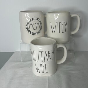 May include: Three white ceramic mugs with black lettering. One mug features the word MOM inside a scalloped circle, another displays the word WIFEY, and the third mug shows the words MILITARY WIFE. Each mug has a handle and a smooth finish.