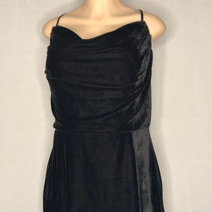 May include: A black velvet dress featuring a cowl neckline and delicate spaghetti straps. The dress has a fitted bodice and a flowing skirt, creating an elegant silhouette. The fabric appears soft and drapes gracefully, suitable for formal events.