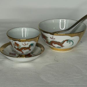 Könnte beinhalten: Eine weiße Porzellantasse auf passender Untertasse und eine größere Schale mit silbernem Löffel, alle mit handgemaltem orange-grünem Drachenmotiv und goldfarbenen Rändern. Das Set zeigt detaillierte Drachenverzierungen auf den Außenseiten.