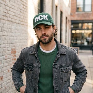 May include: A green baseball cap with the white letters "ACK" is worn. The person is wearing a green sweater and a gray denim jacket. The background is a brick wall and a street.