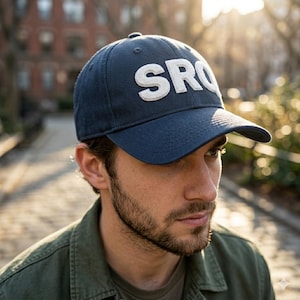 May include: A navy blue baseball cap with the letters "SRQ" in white, textured lettering. The cap has a curved brim and is made of a soft, durable fabric. The hat is a classic style, suitable for everyday wear.