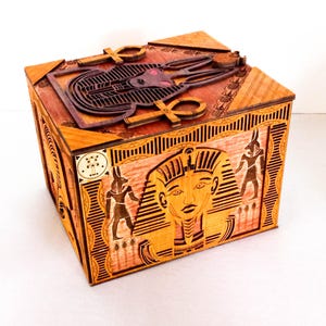 May include: A wooden box with an intricate design featuring Egyptian gods and symbols. The box is decorated with carvings of Anubis, the god of the dead, and the Eye of Horus. The lid of the box features a carving of the goddess Bastet, the goddess of protection.