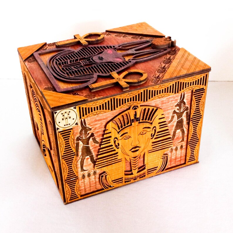 May include: A wooden box with an intricate design featuring Egyptian gods and symbols. The box is decorated with carvings of Anubis, the god of the dead, and the Eye of Horus. The lid of the box features a carving of the goddess Bastet, the goddess of protection.