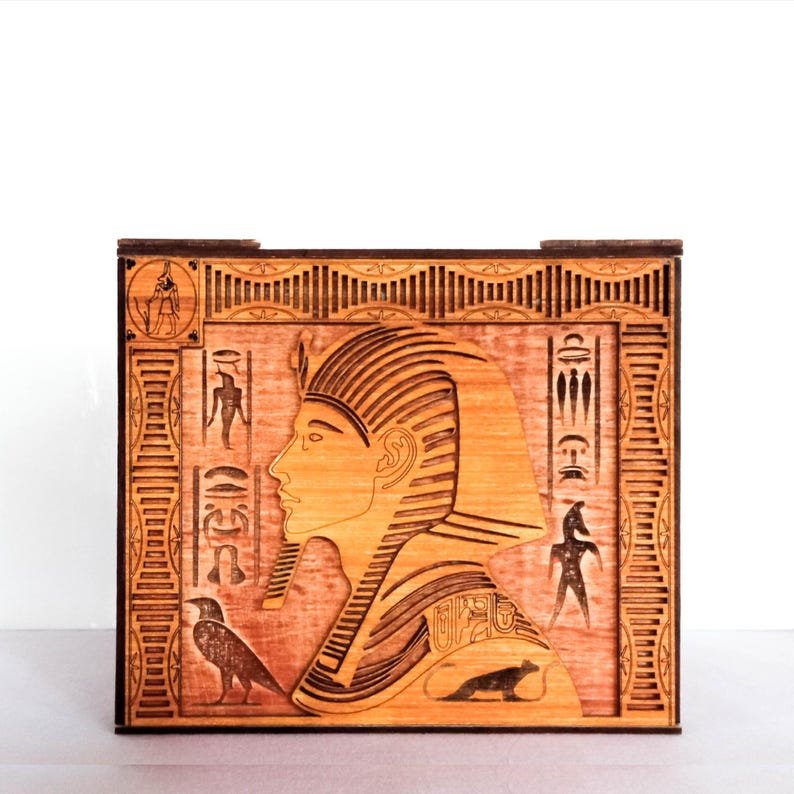 May include: A wooden box with a laser-cut design of an Egyptian pharaoh's profile, hieroglyphics, and birds. The box is stained in shades of brown and has a decorative border.
