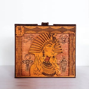 May include: A wooden box with a laser-cut design of an Egyptian queen wearing a crown and jewelry. The box is decorated with traditional Egyptian symbols, including the Eye of Horus, the Ankh, and the Scarab beetle.