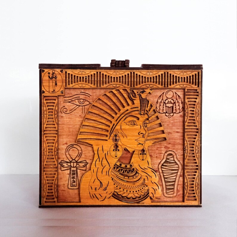 May include: A wooden box with a laser-cut design of an Egyptian queen wearing a crown and jewelry. The box is decorated with traditional Egyptian symbols, including the Eye of Horus, the Ankh, and the Scarab beetle.