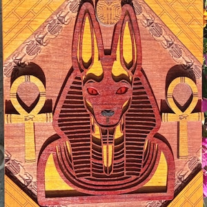 May include: A wooden wall art piece featuring a red and gold Anubis, an Egyptian god, with a scarab beetle above its head. The Anubis is depicted with a human body and a jackal head. The piece is surrounded by a decorative border.