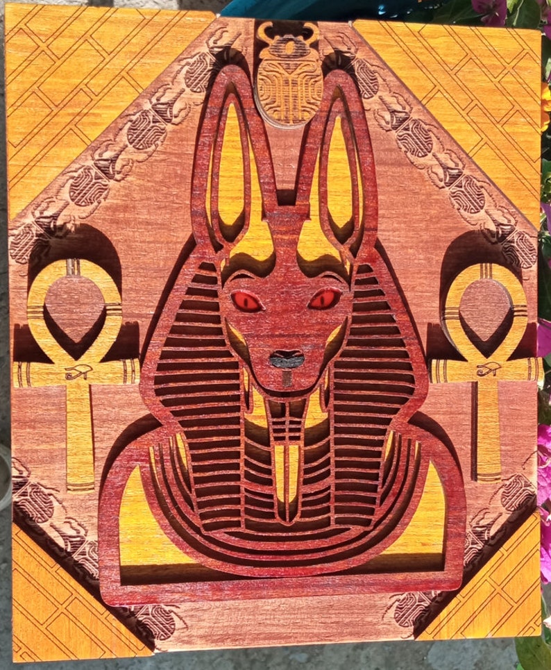 May include: A wooden wall art piece featuring a red and gold Anubis, an Egyptian god, with a scarab beetle above its head. The Anubis is depicted with a human body and a jackal head. The piece is surrounded by a decorative border.