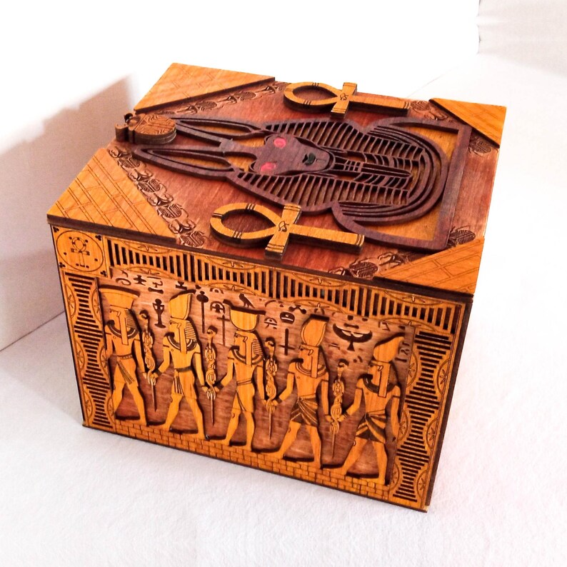 May include: A wooden box with intricate carvings of Egyptian deities and symbols. The top features a detailed carving of Anubis, the jackal-headed god of the dead, with the Ankh symbol on either side. The sides of the box are carved with hieroglyphics and images of Egyptian figures.