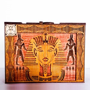 May include: A wooden box with an intricate design featuring an Egyptian pharaoh's face, flanked by two Anubis figures. The box has a red-brown stain and a decorative border.