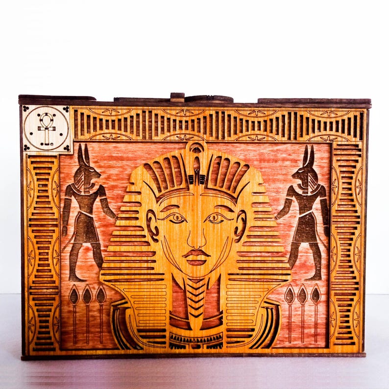 May include: A wooden box with an intricate design featuring an Egyptian pharaoh's face, flanked by two Anubis figures. The box has a red-brown stain and a decorative border.