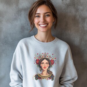 May include: A gray sweatshirt with a colorful illustration of a woman wearing a crown of flowers and a floral patterned dress. The woman has long brown hair and blue eyes.