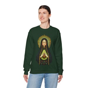 May include: A dark green sweatshirt with a graphic print of a woman holding an avocado. The woman is wearing a gold halo and a blue robe. The avocado is green and has a brown pit.