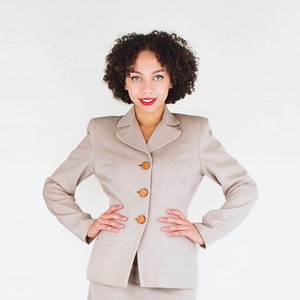 May include: A woman wearing a light beige skirt suit with brown buttons. She is standing with her hands on her hips and is wearing red shoes.