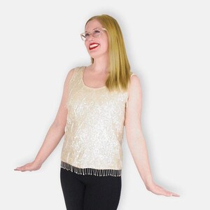 Puede incluir: Una mujer con un top blanco de lentejuelas con un dobladillo de flecos negros, pantalones negros y zapatos blancos. Está sonriendo y mirando hacia arriba.
