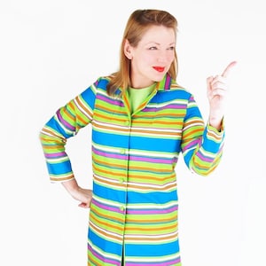 Puede incluir: Una mujer lleva un vestido de manga larga a rayas con un esquema de color verde, azul, morado y naranja. El vestido tiene un frente abotonado y un cuello. Está de pie con la mano derecha levantada, apuntando hacia la derecha.