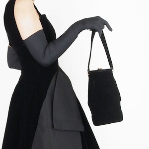 May include: A woman wearing a black velvet dress with a black handbag and long black gloves. She is standing in front of a white background.