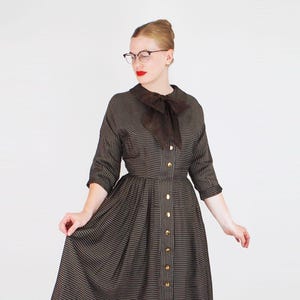 Può includere: Un abito vintage a righe marroni e nere con bottoni sul davanti e un fiocco marrone sul colletto. L'abito ha maniche a tre quarti e una gonna svasata. L'abito è al ginocchio.