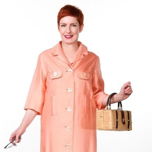 May include: A peach-colored, knee-length coat with short sleeves and white buttons. The coat is paired with black pants and black shoes. A small, woven handbag with a brown handle is held in the right hand. The person is also holding eyeglasses.