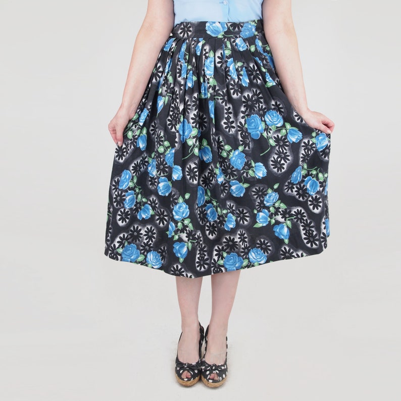 May include: A black midi skirt with a floral pattern featuring blue roses and green leaves. The skirt has a pleated design and is paired with black and white polka dot shoes. The skirt is knee-length.